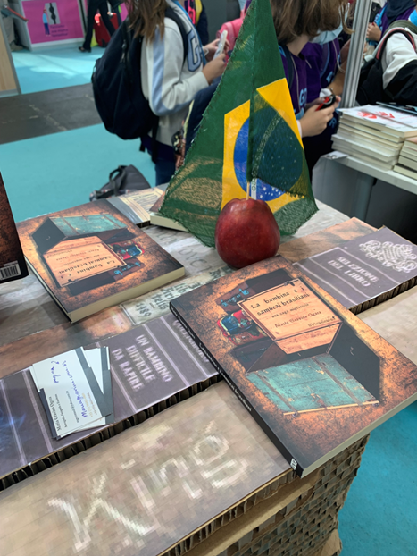 Mesa de autógrafos com livros de Maria Gravina Ogata, bandeira do Brasil e público ao fundo